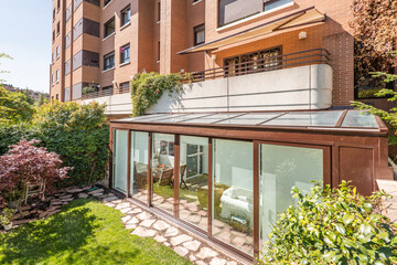 Fototapeta na wymiar Brown aluminum and glass covered terrace in the lower part of a residential apartment building with sliding doors, decorative trees and a cobbled path