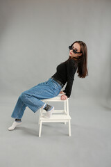 beautiful young woman on a gray background in black sunglasses. sit on a white chair. poses for a photo shoot