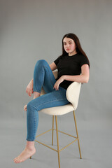 stylish photo shoot beautiful model with black hair. girl in jeans and black turtleneck on a gray background. girl sitting on a white chair and posing for a photo