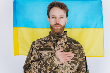 Ukrainian patriot soldier in military uniform holds a hand on a heart against the background of a yellow and blue flag