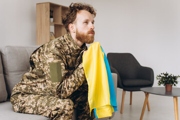 Portrait of an emotional young Ukrainian patriot soldier in military uniform sitting on the office on the couch holding a yellow and blue flag.