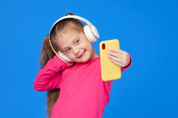 A cute girl in a bright pink sweater taking a selfie on a mobile phone camera and listens to music in wireless headphones. The concept of children's applications on gadgets