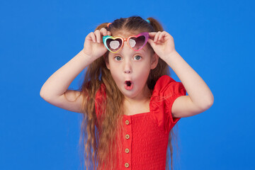 Portrait of surprised cute little toddler girl in the heart shape sunglasses. Child with open mouth having fun isolated over blue background. Looking at camera. Wow funny face