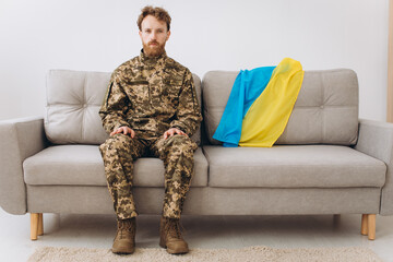Portrait of an emotional young Ukrainian patriot soldier in military uniform sitting on the office on a sofa with a yellow and blue flag.