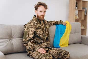 Portrait of an emotional young Ukrainian patriot soldier in military uniform sitting on the office on a sofa with a yellow and blue flag.
