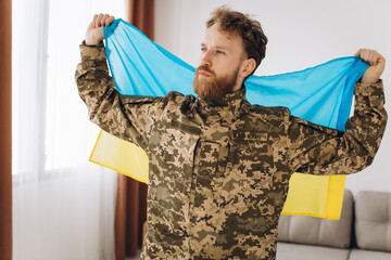 Portrait of an emotional young bearded Ukrainian patriot soldier in military uniform holding a flag...