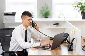 young man in a white shirt with a black tie at the desk at the bright office working on a laptop