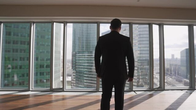 Rear view of a young successful businessman walking in an office and looking through a full length window at a cityscape with skyscrapers. Businessman looks out the window in the city.