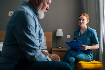 Obraz premium Female doctor writing down diagnosis and symptoms in medical history on clipboard, talking to sick senior male patient sitting on bed at home. Elderly man getting medical consultation in hospital.