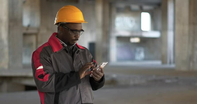 African american builder at construction site uses smartphone. Workman in yellow hard hat and goggles stands at project site and works with smartphone. Cinema 4K 60fps zoom video