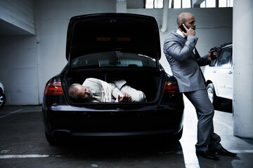 Hostage to the mob. A frightened businessman lying in the trunk of a car while his armed captor talks on a cellphone. - obrazy, fototapety, plakaty