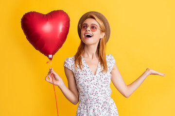 happy girl in straw hat and sunglasses hold love heart balloon on yellow background