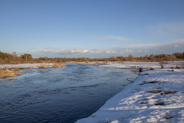 A picturesque landscape, early spring, a river with snow-covered banks, dry grass and bushes. March sunny day by the river. The first thaws, the snow is melting.