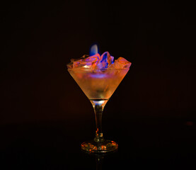 Cocktail glass on a dark background, in which ice burns with a blue flame. Orange ice burns in a glass with blue reflections.