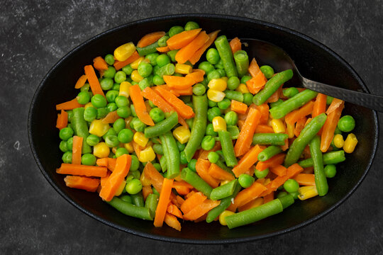 Mixed vegetables, peas, carrots, green beans and corn, in a black dish.. On a black stone background