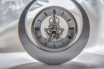 A modern analog clock with a sculpted silver body and see through face showing internal clock gear mechanism