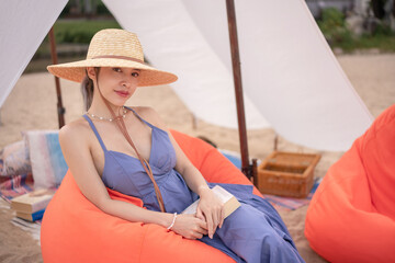 Woman wearing blue dress and straw hat sitting on orange beanbag near white tent on a beach.