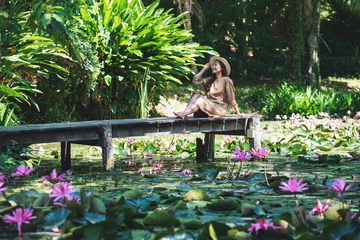 Fotobehang Portrait image of a beautiful young asian woman sitting on wooden bridge in a pond with pink lotus flowers © Farknot Architect