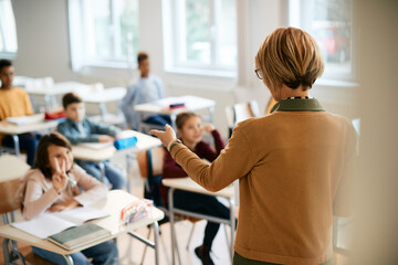 Rear view of elementary school teacher points at one of her students who is raising arm to answer a...