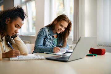 Happy high school student writes while e-learning on laptop with her classmate in the classroom.