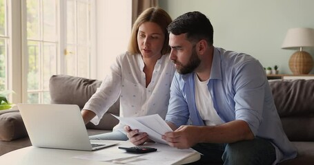 Attractive millennial couple sit on sofa do paperwork, reviewing household utility bills, sorting out documents, manage family finances, planning investments, pay taxes online via e-bank app on laptop