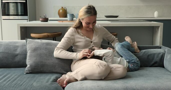 Kind mommy babysitter sit on sofa play cuddle tickle adorable little daughter girl preschooler caress beloved kid laugh have fun together. Positive energetic mom child girl mess enjoy funny pastime