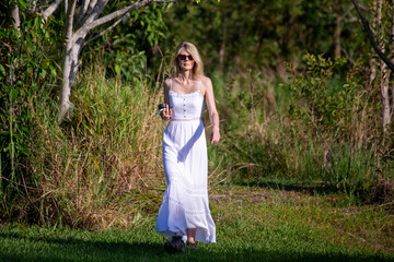 Blonde Woman in White Dress Holding Phone and Camera Lens Emerging from Forest Looking at Us