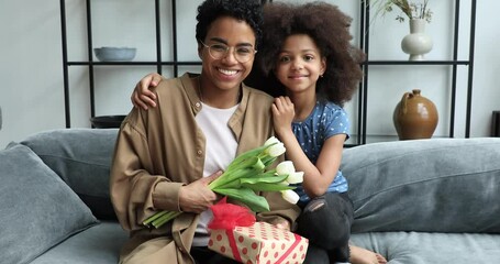Excited Black mother sit on couch embraced by beloved child girl look at camera with grateful smile hold gift box flowers. Portrait happy single mom get present tulips on Women Day from tween daughter