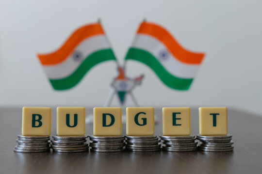 Letter blocks arranged as 'BUDGET' on pile of coins and blur Indian flag in the background. Main focus is on letter blocks and coins. Flag is purposely blur.