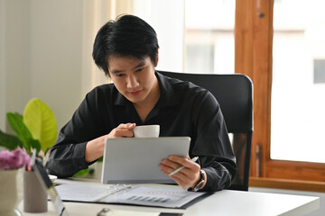 A portrait of a young Asian man sitting in the office holding a cup of coffee and working on data and documents and a laptop on the table, for business and  technology concept.
