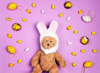 Easter background with a funny toy bear with rabbit ears and colored eggs on purple.