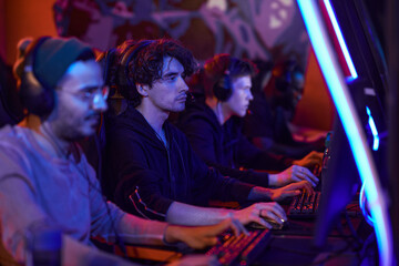 Obraz premium Group of concentrated young men in headsets sitting in row and playing video games in esports bar