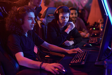 Group of concentrated team members looking at computer monitor and assisting guy to win game in neon-illuminated room
