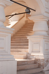 A white stone marble bright classic staircase are leading up with a curve and pillars with an ornament. Sunrays are passing through and lit the stairs