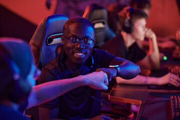 Smiling young Black man in glasses sitting into computer chair and giving fist bump to team member...