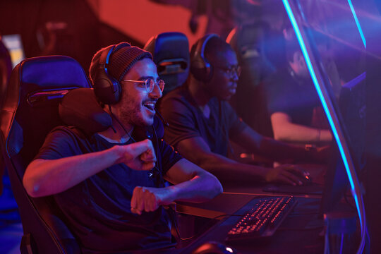 Happy Young Middle Eastern Professional Gamer In Headphones And Eyeglasses Dancing In Front Of Computer While Completing Video Game Misson