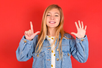 blonde little kid girl wearing denim jacket over red background showing and pointing up with fingers number seven while smiling confident and happy.