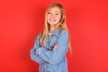 blonde little kid girl wearing denim jacket over red background happy face smiling with crossed arms looking at the camera. Positive person.