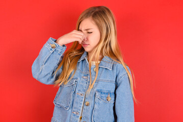 blonde little kid girl wearing denim jacket over red background smelling something stinky and disgusting, intolerable smell, holding breath with fingers on nose. Bad smell