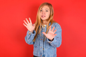blonde little kid girl wearing denim jacket over red background afraid and terrified with fear expression stop gesture with hands, shouting in shock. Panic concept.