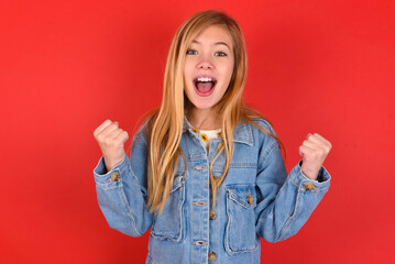 blonde little kid girl wearing denim jacket over red background celebrating surprised and amazed...