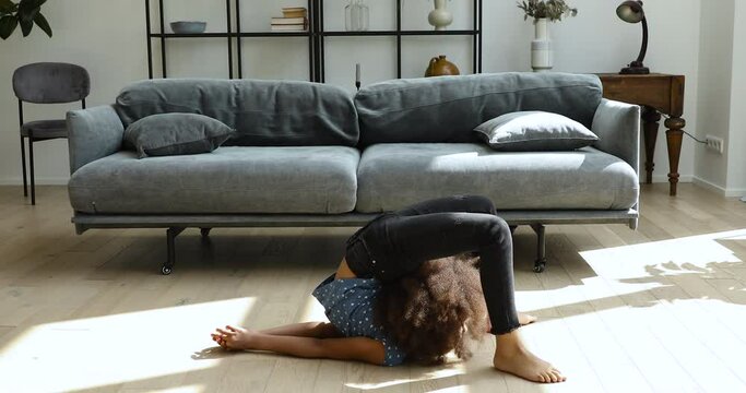Flexible preteen age Afro American girl do gymnastic exercise at living room on warm floor with underheating. Sporty teen kid engaged in physical training practice yoga acrobatics stretch back muscles