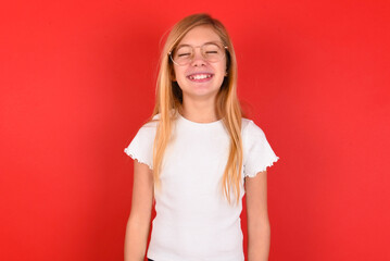 Positive blonde little kid girl wearing white t-shirt over red background with overjoyed expression...