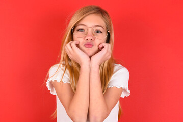 blonde little kid girl wearing white t-shirt over red background with surprised expression keeps hands under chin keeps lips folded makes funny grimace