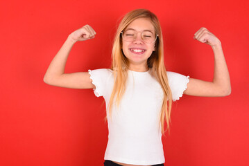 Strong powerful blonde little kid girl wearing white t-shirt over red background toothy smile,...