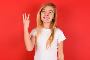 Obraz na płótnie Canvas blonde little kid girl wearing white t-shirt over red background smiling and looking friendly, showing number three or third with hand forward, counting down