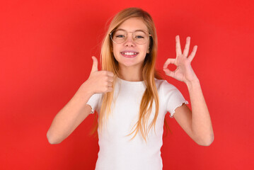 blonde little kid girl wearing white t-shirt over red background smiling and looking happy, carefree and positive, gesturing victory or peace with one hand