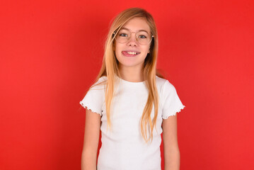 Funny blonde little kid girl wearing white t-shirt over red background makes grimace and crosses eyes plays fool has fun alone sticks out tongue.