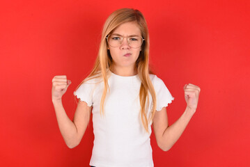 Fototapeta premium Irritated blonde little kid girl wearing white t-shirt over red background blows cheeks with anger and raises clenched fists expresses rage and aggressive emotions. Furious model