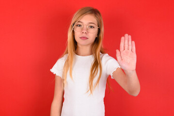 Fototapeta premium blonde little kid girl wearing white t-shirt over red background shows stop sign prohibition symbol keeps palm forward to camera with strict expression